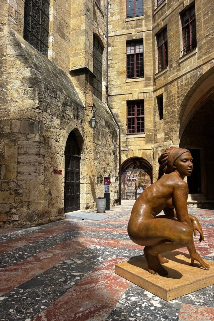 Statue of a crouching woman in the courtyard of the Palais Neuf, surrounded by historic stone buildings with arched doorways and windows.