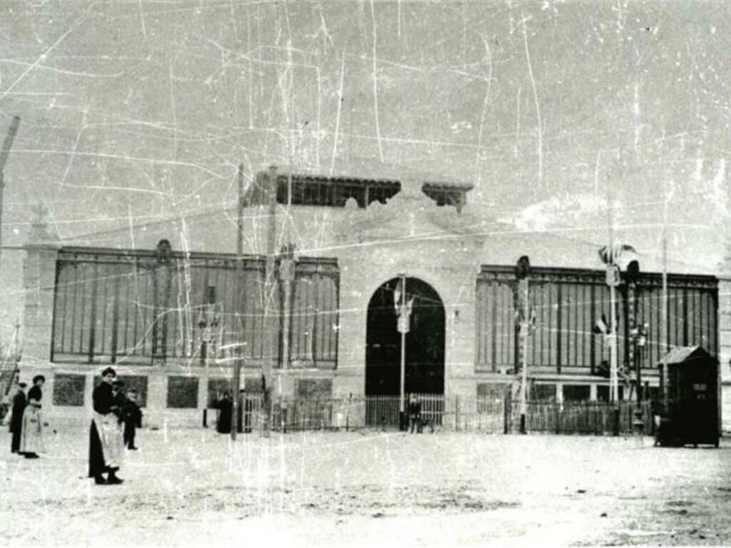 A grainy, black-and-white historical photo showing the exterior of Narbonne Market in its early days. The building has large windows, and people dressed in period clothing stand outside, giving a glimpse into the market's past.