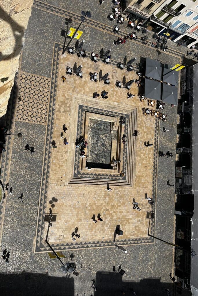 Bird's-eye view of Via Domitia from the Donjon in Narbonne, showing a patterned square with people walking and outdoor seating areas.