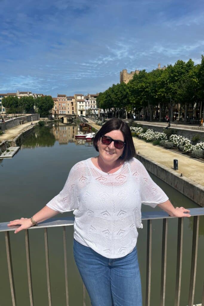A woman in sunglasses and a white knit top smiles while standing on a bridge in front of a calm canal with colorful buildings and trees in the background. The sunny day highlights the picturesque cityscape of Narbonne.