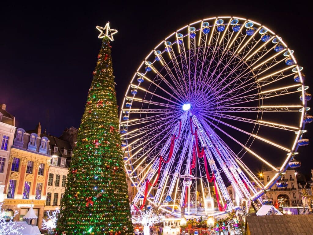 A colorful night scene at Lille Christmas Market, featuring a large Ferris wheel and a towering Christmas tree, both brightly lit against the night sky, surrounded by festive decorations and historic buildings.