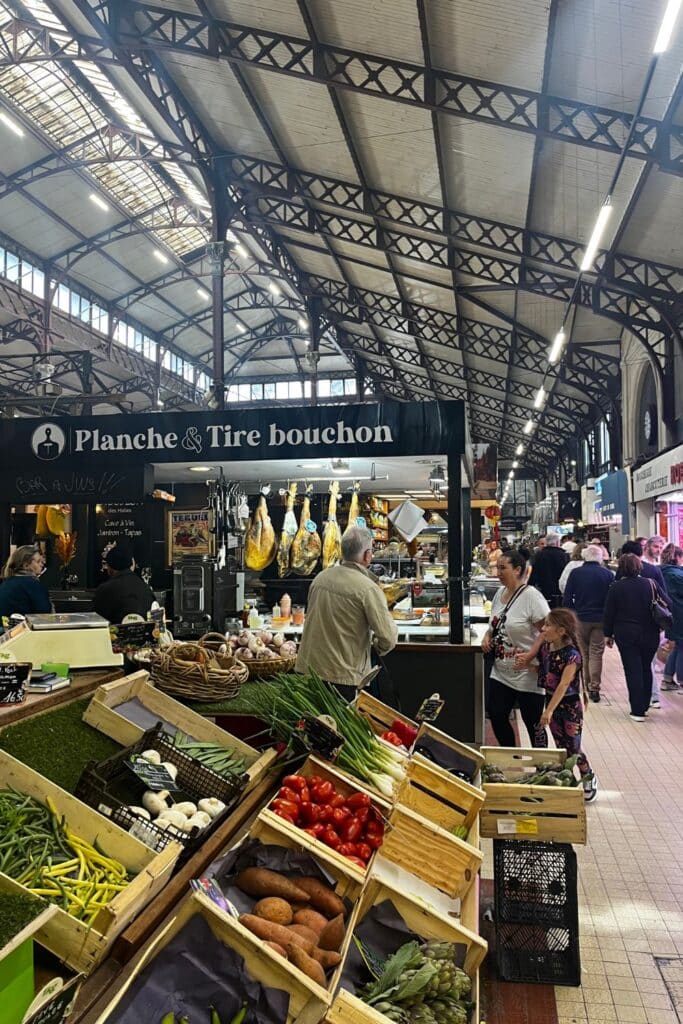 An indoor market scene at "Planche & Tire bouchon," showcasing fresh produce like tomatoes, onions, and green beans in wooden crates. In the background, cured meats hang from the stall as people walk through the busy market.
