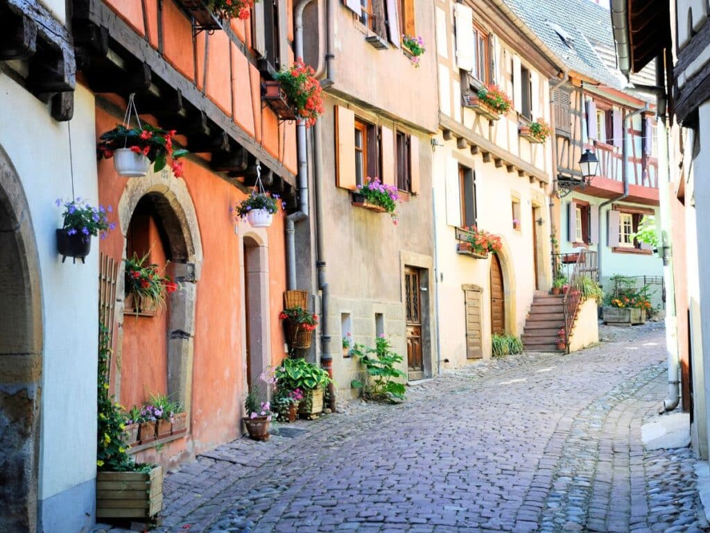 A cozy Alsatian village street lined with pastel-colored, timber-framed houses. Vibrant flowers hang from balconies and windows, with the cobblestone street leading to a hidden corner, evoking the region’s storybook charm.