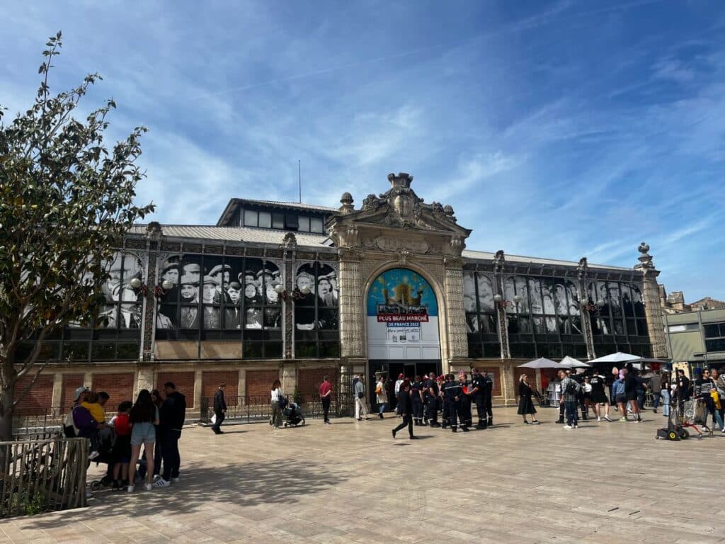 A modern-day view of Narbonne Market with its Belle Époque architecture, featuring large windows decorated with vintage black-and-white photos of people. Visitors gather outside under the blue sky, while vendors set up stalls on the plaza.