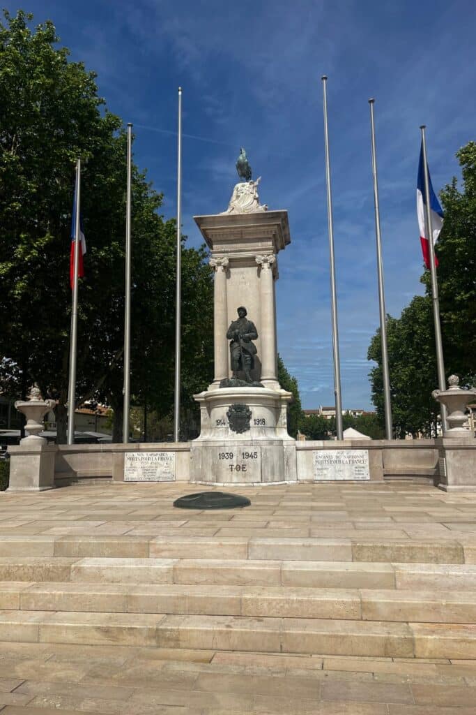 A World War memorial statue in Narbonne, France, commemorating soldiers from the 1914-1918 and 1939-1945 wars. The stone monument is adorned with French flags, with intricate sculptures and inscriptions honoring the fallen.