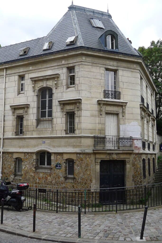 An elegant, historic building with a light beige stone exterior and a sloped grey slate roof. The structure displays French architectural style with barred windows, wrought iron railings, and a corner entrance bordered by cobblestone streets and metal railings.