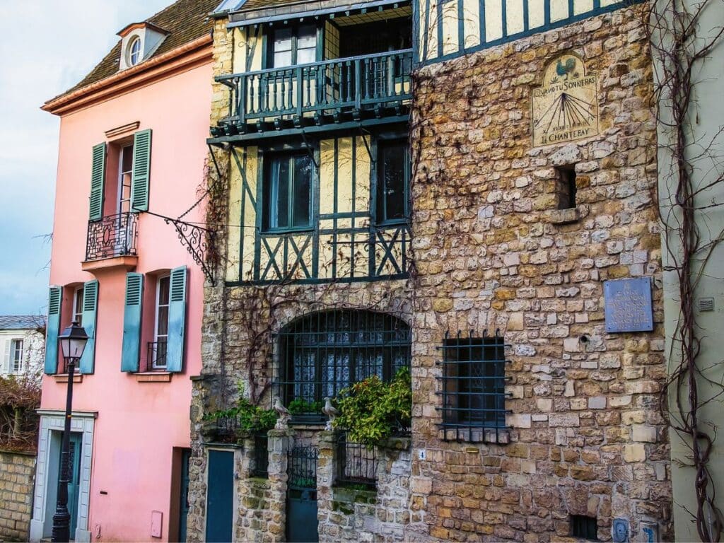 A charming street in Montmartre featuring a rustic building with a stone façade. It has a distinct sundial with the inscription "Quand tu sonneras, je chanteray." To the left, a pastel pink building with green shutters and wrought iron balcony adds a pop of color.