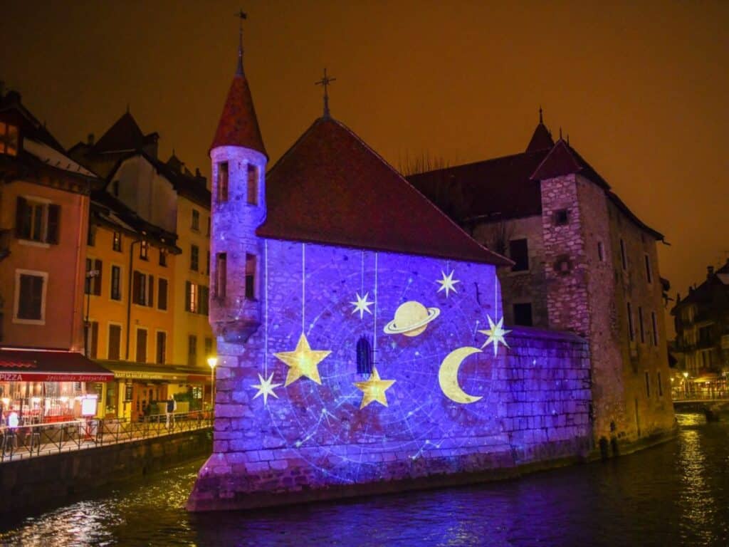 A nighttime scene of an old stone building in Annecy, France, with projections of stars, a crescent moon, and planets illuminating its facade. The building is surrounded by a softly lit, tranquil canal and charming, brightly colored Alpine-style houses.