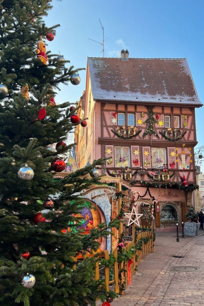 A large decorated Christmas tree with ornaments like silver baubles and gingerbread men stands beside a pastel-colored half-timbered house adorned with stars, garlands, and holiday decorations in Colmar, France.