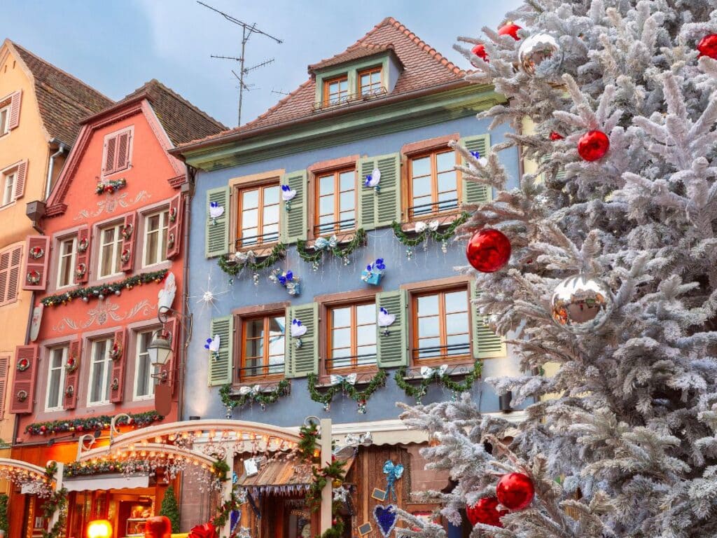 A frosted Christmas tree with red and silver baubles stands beside colorful half-timbered houses decorated with festive garlands and ornaments in a Christmas market setting in Colmar, France.
