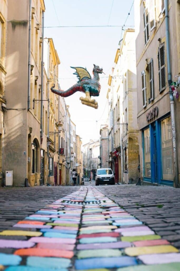 A colorful dragon sculpture, representing the local legend of the Graoully, hovers between the historic buildings of Metz. Below it, a colorful brick road leads into the distance of the narrow street.