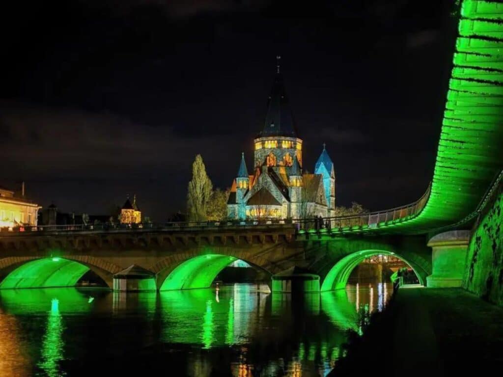 A nighttime view of a bridge illuminated in vibrant green light, reflecting off the water below. In the background, a beautifully lit church with spires stands prominently, surrounded by the serene river and distant trees.