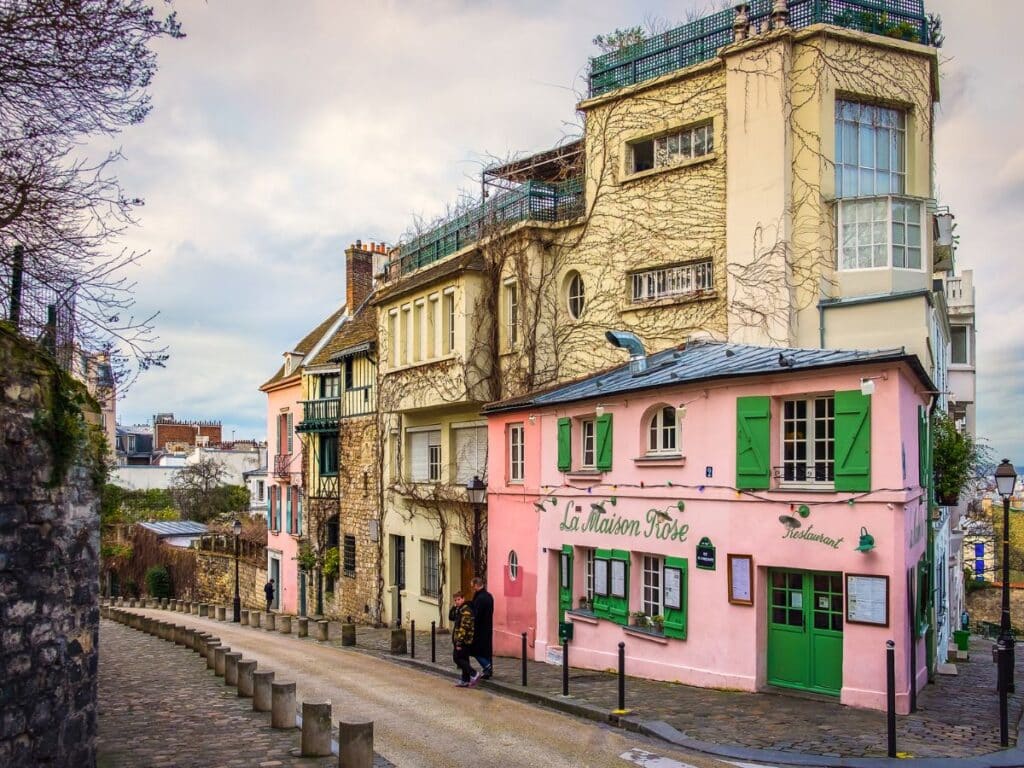 A picturesque scene of the famous "La Maison Rose" café in Montmartre, Paris. The café is painted in pink with green shutters and is situated on a charming, winding cobblestone street lined with classic Parisian buildings covered in vines.
