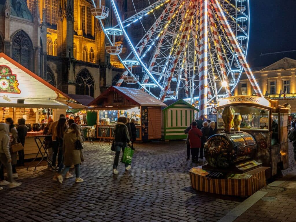 A lively evening at a Christmas market with people walking among the brightly lit stalls. A towering Ferris wheel and the illuminated facade of a historic building add to the festive atmosphere.