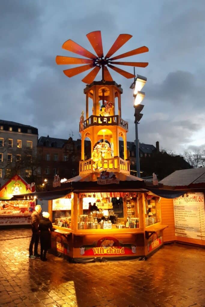 A wooden Christmas pyramid stall lit up in the early evening, its spinning blades adding a whimsical touch. The cozy stall, offering "vin chaud" (hot wine), is set against a backdrop of darkening skies and festive holiday markets.