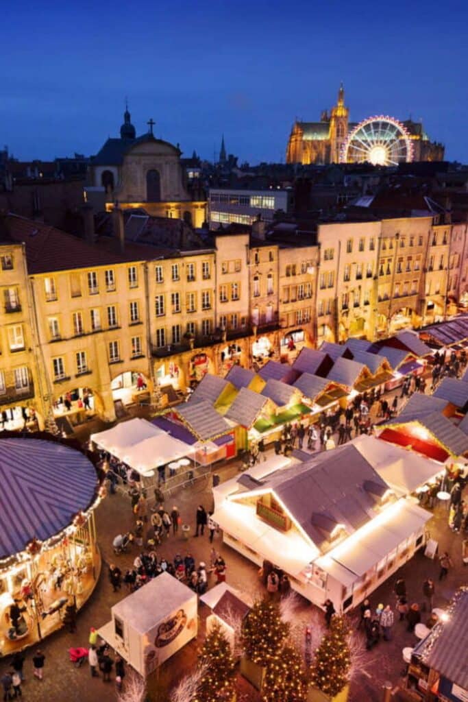 An aerial view of the Metz Christmas market in the evening, showcasing rows of colorful stalls beneath glowing streetlights. The Cathedral of Metz and a brightly lit Ferris wheel dominate the background, creating a festive atmosphere in the heart of the city.