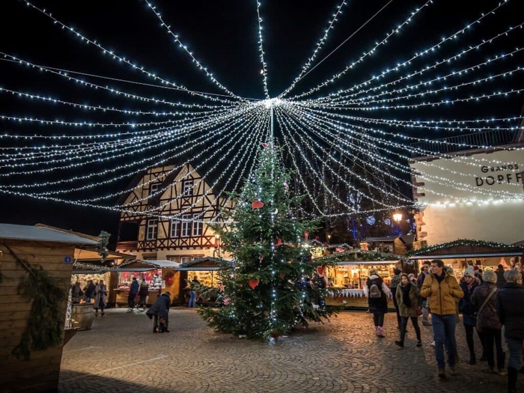 A large Christmas tree stands in the center of a town square, with bright string lights radiating from the top, creating a magical atmosphere as market-goers explore the surrounding stalls