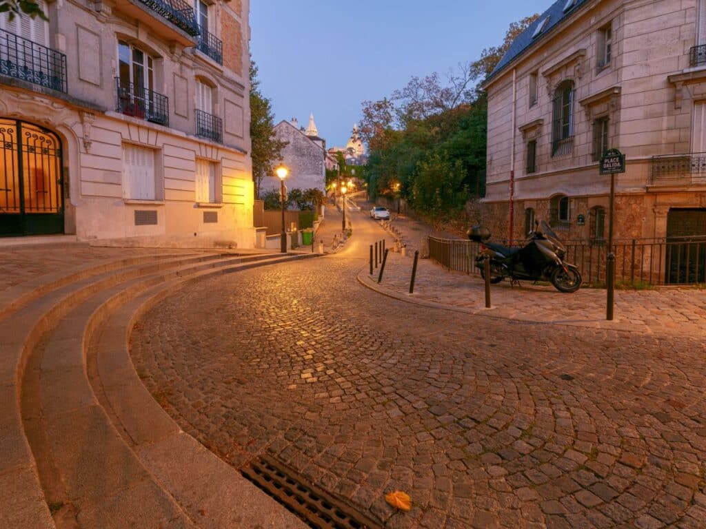 A scenic, winding street in Montmartre with a gentle incline lined by traditional Parisian buildings in soft pastel colors. A few pedestrians stroll along the cobblestone path, capturing the historic and artistic essence of the neighborhood.