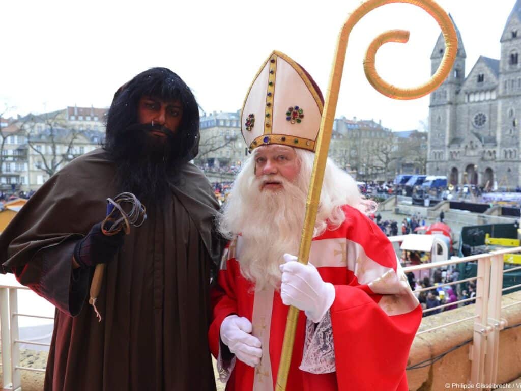 A photo of two men dressed as Saint Nicholas and his companion, the dark-robed Père Fouettard. Saint Nicholas holds a golden staff, while Père Fouettard carries a bundle of sticks, standing against a backdrop of a festive public gathering.