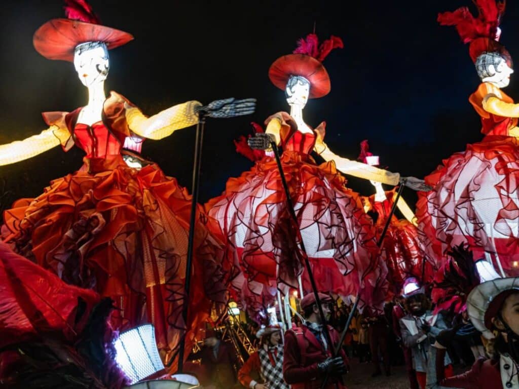 A parade of large, glowing puppets in red dresses and wide-brimmed hats, carried by performers in costume. The figures stand tall, illuminated by the lanterns inside them, surrounded by a dark night sky that highlights the brightness of the display.