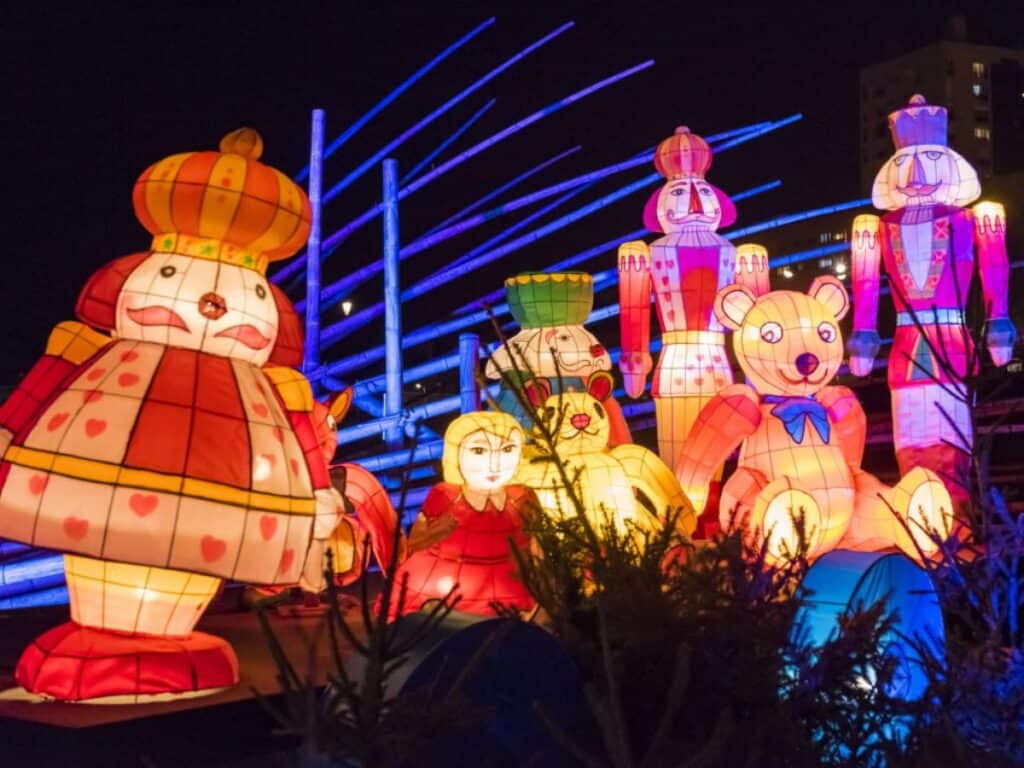 A whimsical display of large, colorful lanterns shaped like Nutcracker figures and stuffed toys. The lanterns glow in vibrant hues of red, yellow, and blue, set against a night sky, giving the scene a magical, fairytale-like atmosphere.