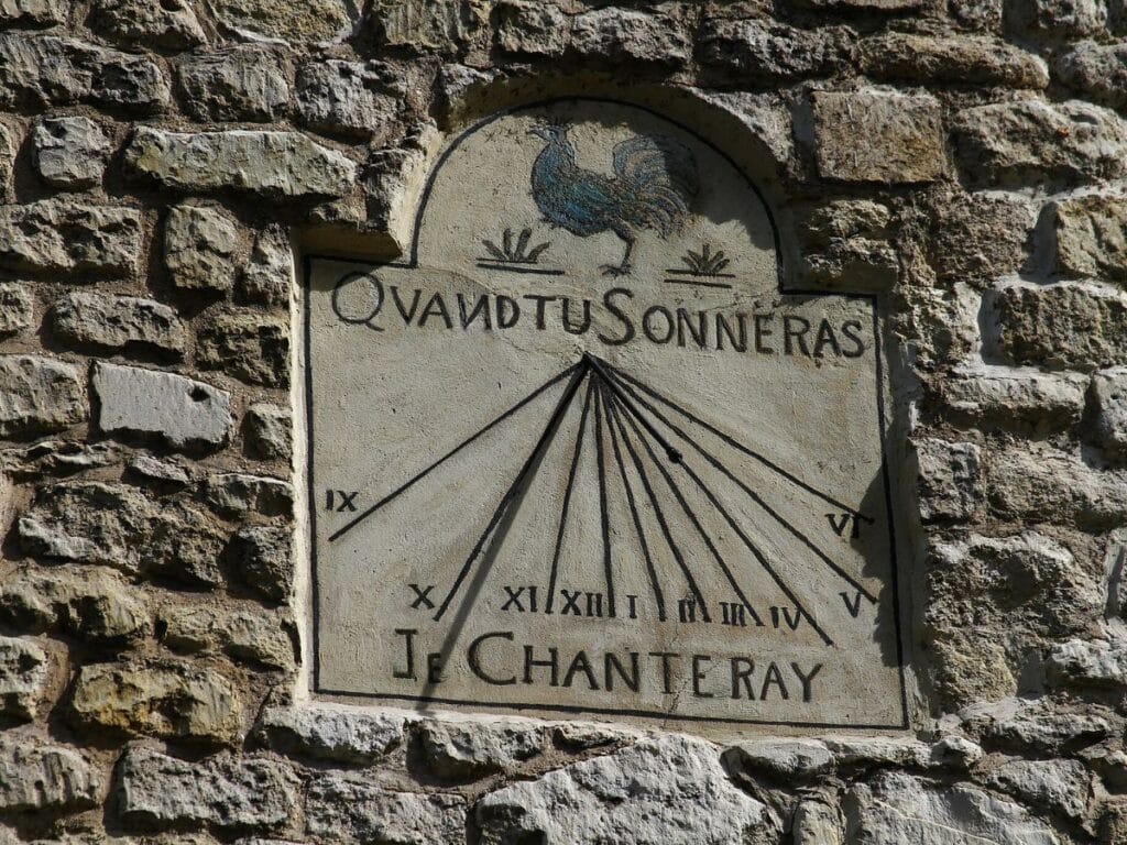 A close-up of a stone sundial embedded in a rustic wall at 4 Rue de L’Abreuvoir. The inscription reads, "Quand tu sonneras, je chanteray," surrounded by Roman numerals and the silhouette of a rooster at the top.