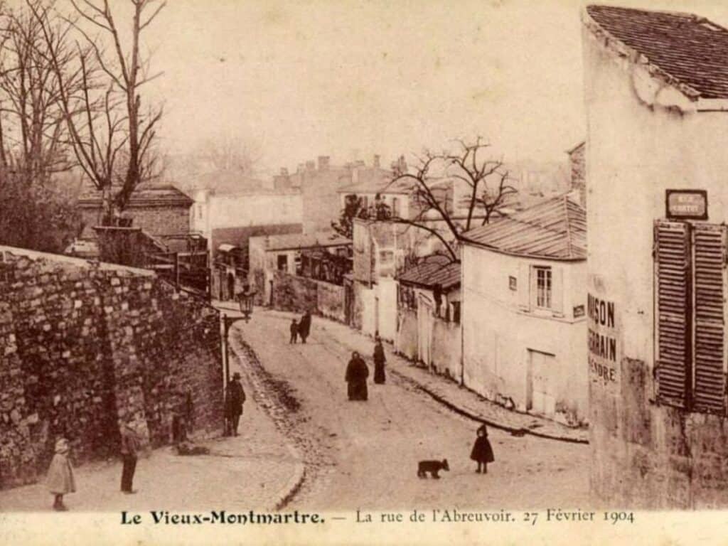 A sepia-toned historical photograph of Rue de L’Abreuvoir from February 27, 1904. It depicts the winding cobblestone street, with a few pedestrians and a dog walking through, lined with stone walls and traditional buildings, giving a glimpse into Paris' past.