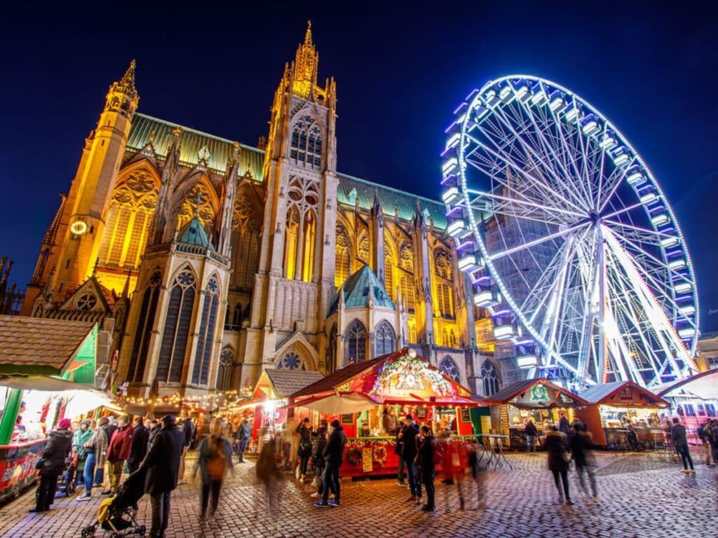 The Metz Cathedral lit up with warm golden light, next to the glowing Ferris wheel, as crowds of visitors stroll through the bustling Christmas market stalls below. The festive energy fills the cobblestone square, illuminated by decorative lights.