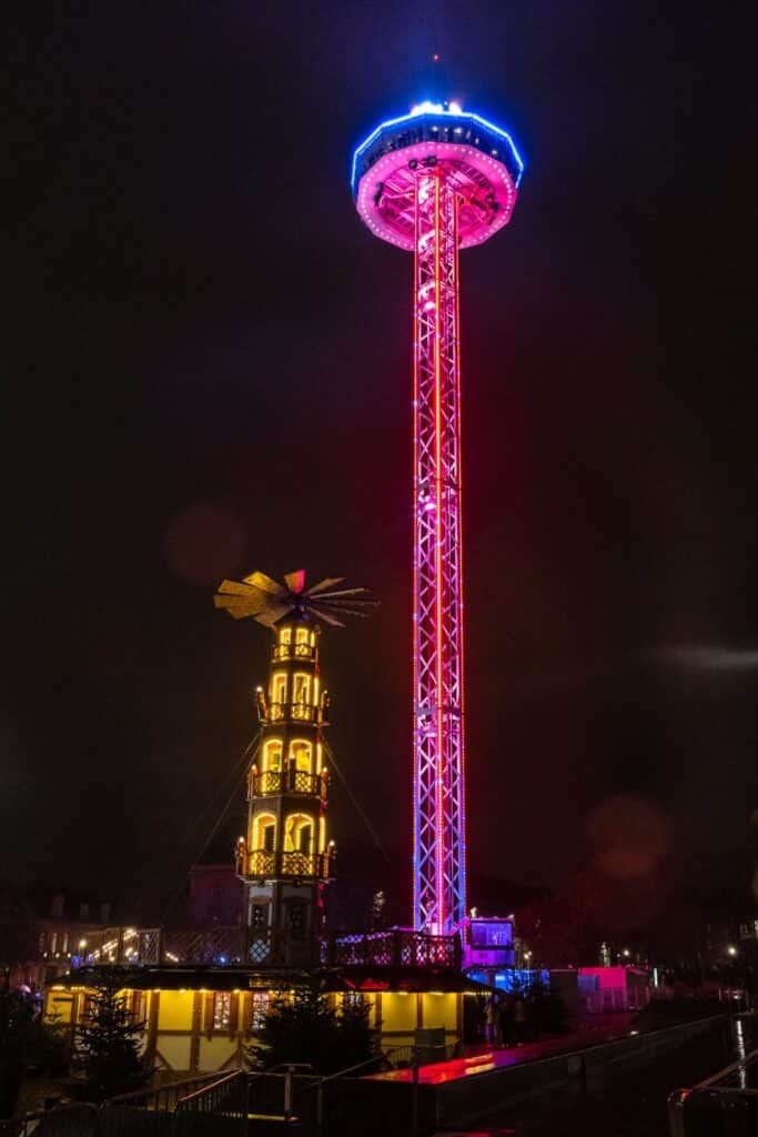 A nighttime skyline of Metz, with the glowing Ferris wheel and illuminated Cathedral of Metz towering above the city’s rooftops. The market lights and bustling crowds below give the city a festive, holiday charm.