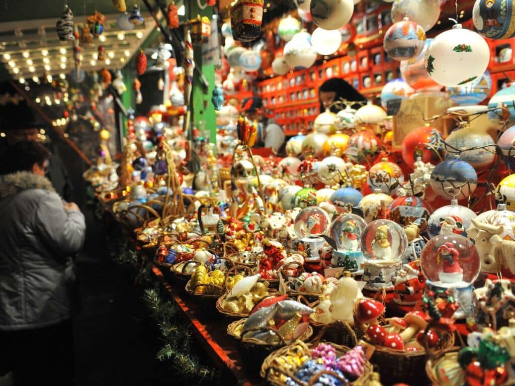 A bustling Christmas market stall filled with an assortment of colorful ornaments, snow globes, and festive decorations. Shoppers browse the vibrant collection under warm string lights, capturing the cozy and lively holiday spirit.