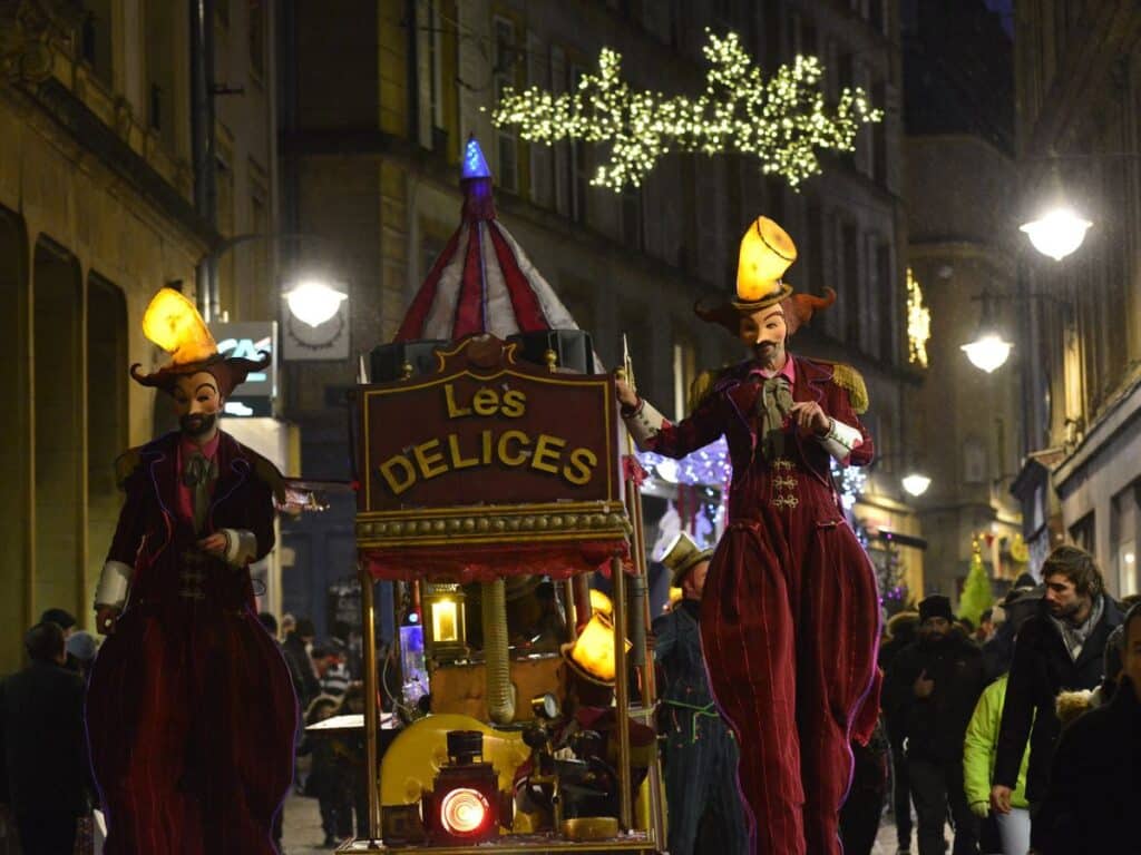 Two costumed performers in extravagant red outfits with illuminated hats stroll through the streets during a night parade. They walk beside a decorated cart labeled "Les Délices," entertaining onlookers as festive lights twinkle overhead.