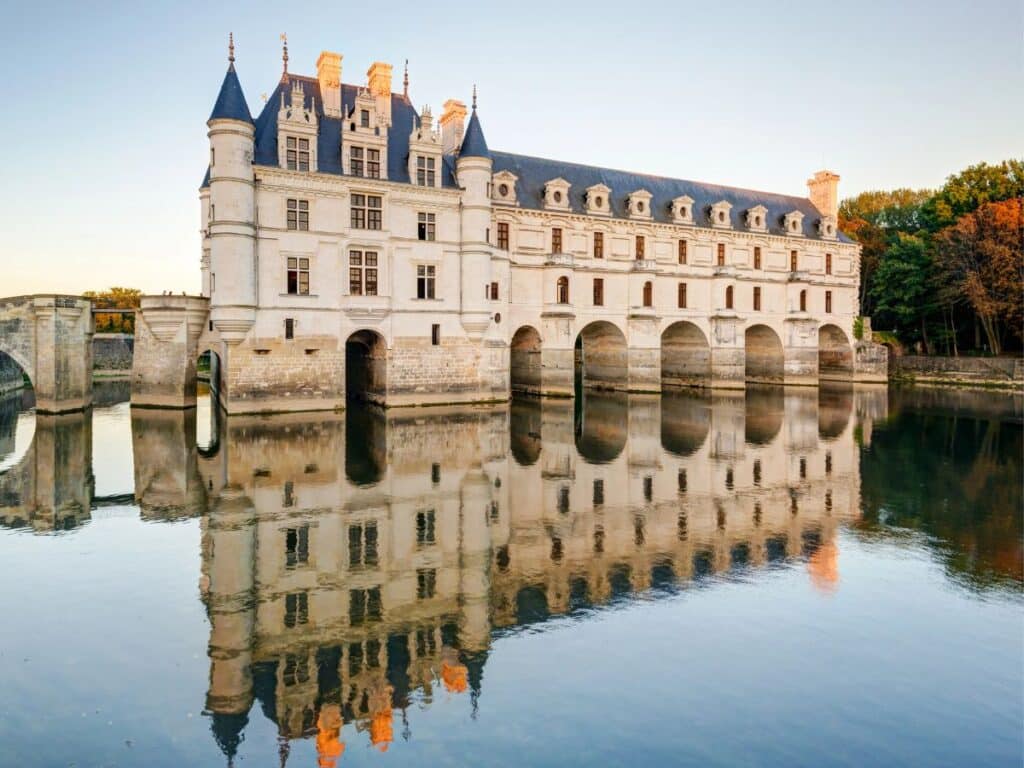 An aerial view of Château de Chenonceau showcasing its iconic arches over the river and its beautifully manicured gardens. The surrounding forests and river reflect the estate's natural beauty.