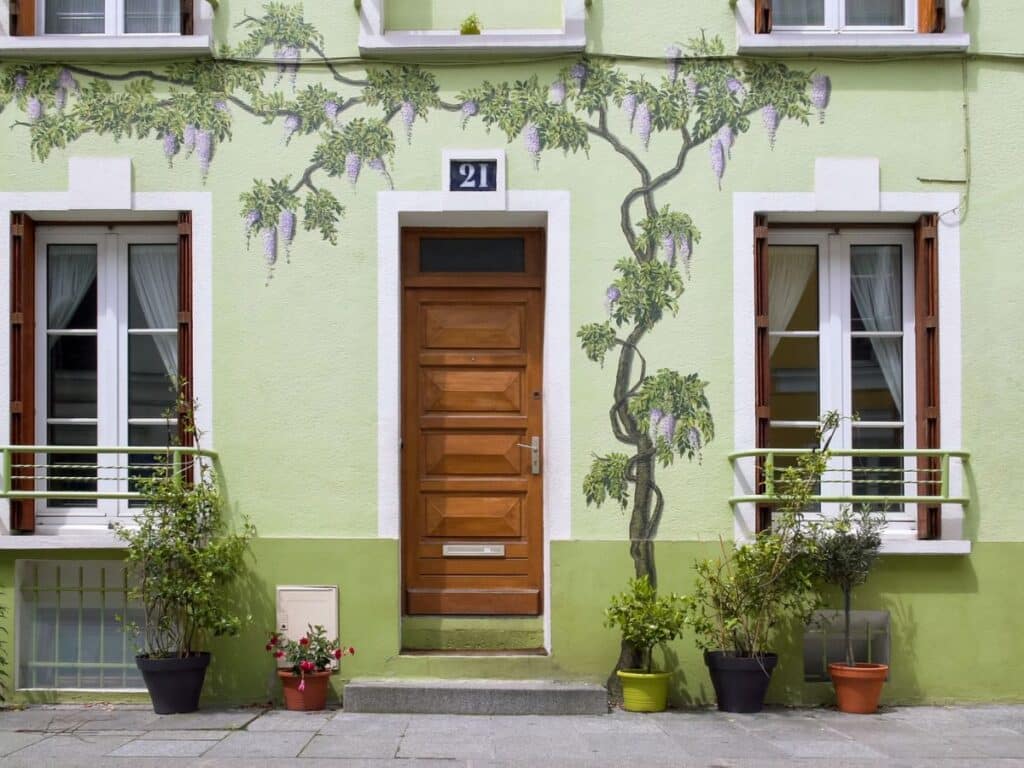 A charming building facade painted in pastel green, adorned with delicate vine murals climbing the wall. The wooden front door is centered, flanked by windows with wooden shutters and potted plants, creating a quaint, welcoming aesthetic.