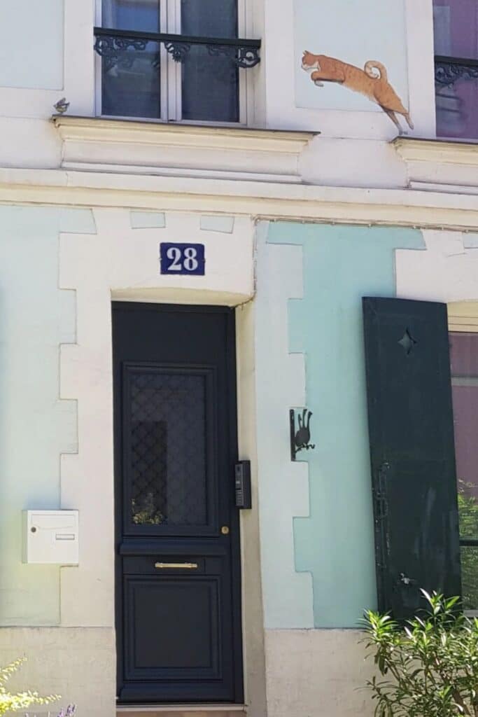 A light pastel blue and cream building with a black door and a mural of a leaping orange cat above. The building has a classic European style, with green shutters on one side and a small mailbox mounted beside the door.
