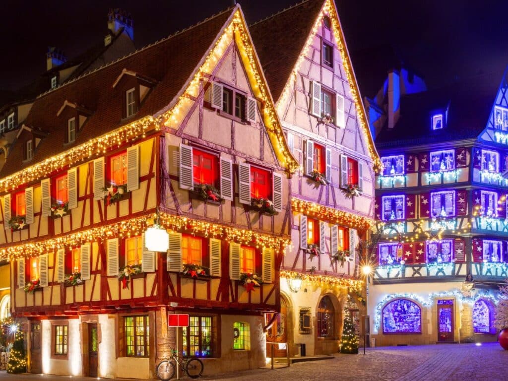 A beautifully lit, traditional half-timbered building in what appears to be a European village during Christmas. The building is adorned with festive string lights, wreaths, and red shutters, creating a warm, holiday atmosphere. In the background, another building is similarly decorated with lights in a blue and purple hue, enhancing the magical ambiance of a winter wonderland scene.