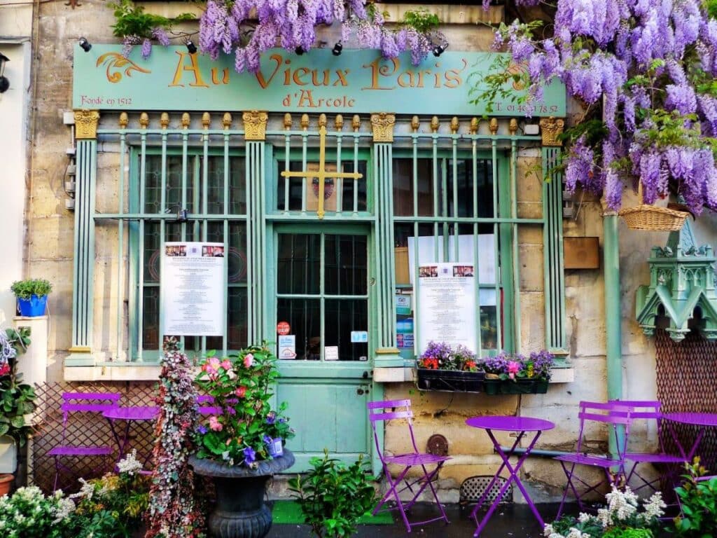 A charming Parisian café, "Au Vieux Paris d'Arcole," covered in blooming wisteria vines. The front of the café is adorned with teal window frames and golden signage. Outside, bright purple tables and chairs are arranged on the cobblestone sidewalk, surrounded by potted plants and vibrant flowers.