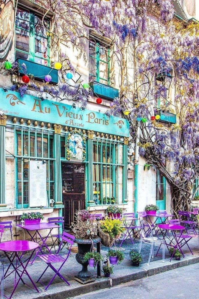 A charming Parisian café, "Au Vieux Paris d'Arcole," covered in blooming wisteria vines. The front of the café is adorned with teal window frames and golden signage. Outside, bright purple tables and chairs are arranged on the cobblestone sidewalk, surrounded by potted plants and vibrant flowers.