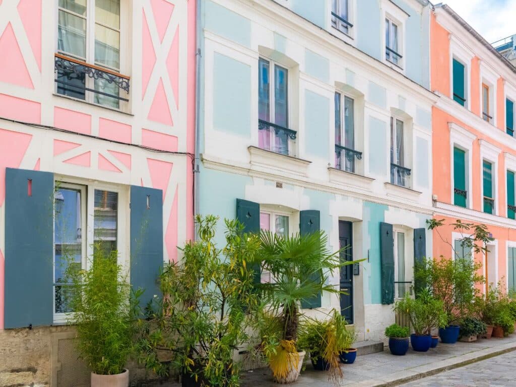 A street scene of vibrant, pastel-colored houses in a row. Each building has distinctive colors with matching shutters, and potted greenery lines the walkway, adding to the lively and artistic ambiance.
