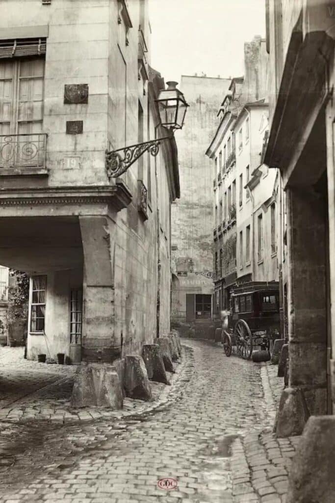 A sepia-toned vintage photograph of a narrow cobblestone street lined with old Parisian buildings. An ornate street lamp and a horse-drawn carriage add to the historic ambiance of the scene.