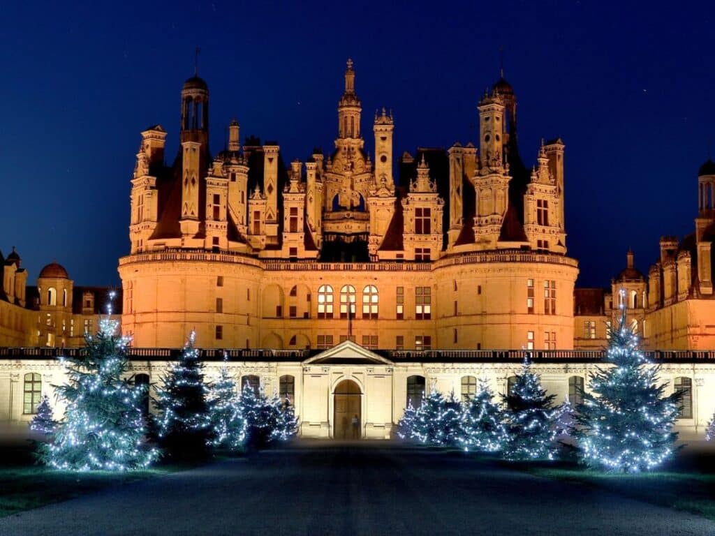 A grand château illuminated at night, with its ornate spires and towers lit up in a golden glow against a deep blue sky. In the foreground, Christmas trees decorated with white string lights are arranged along the pathway, creating a festive and majestic scene. The contrast between the historic architecture and the holiday lights evokes a magical winter atmosphere, characteristic of France during the Christmas season.