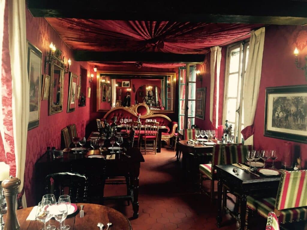 An elegant, warmly lit dining room inside "Au Vieux Paris d'Arcole." The walls are covered in deep red fabric with golden accents, while mirrors and framed artworks decorate the space. Tables are set with wine glasses, plates, and utensils, surrounded by colorful striped chairs.