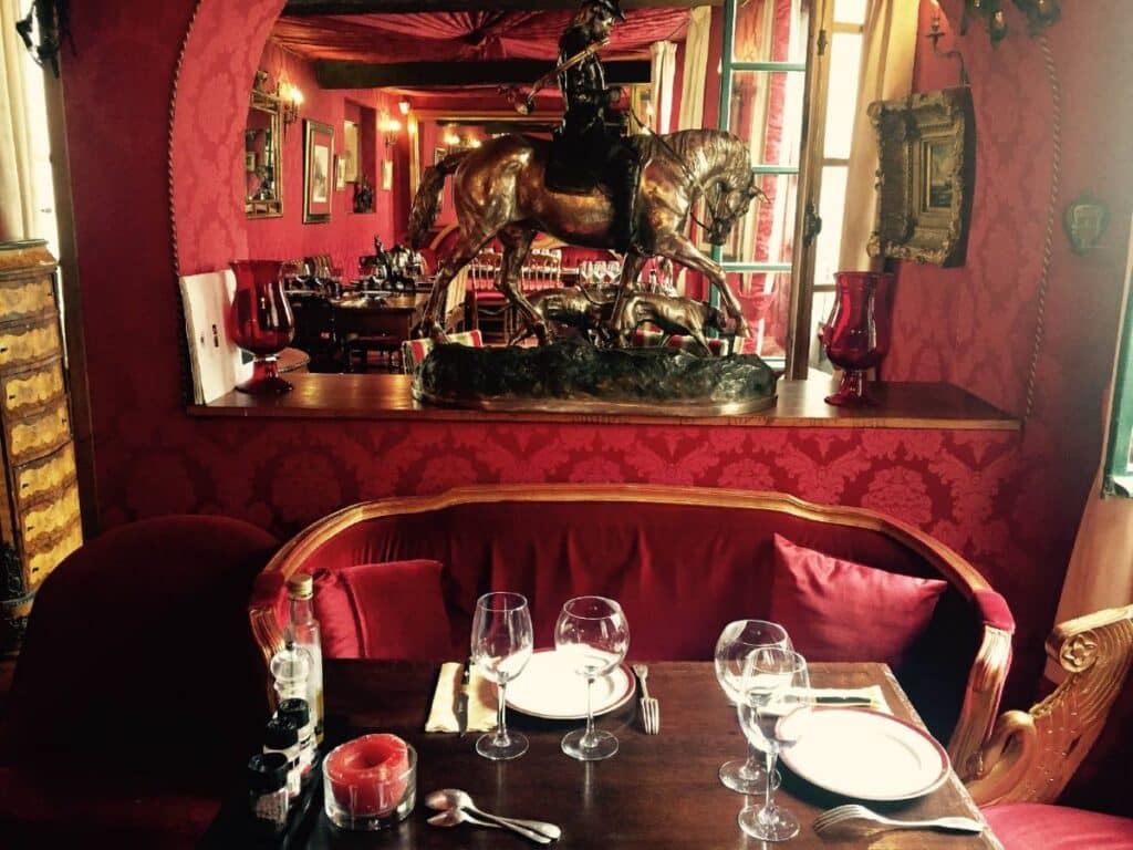 A cozy dining table set for two in "Au Vieux Paris d'Arcole." The room is adorned in rich red decor, with plush seating, red velvet curtains, and a bronze statue of a horse and rider on a shelf in the background. Wine glasses, plates, and cutlery are neatly arranged on the wooden table.
