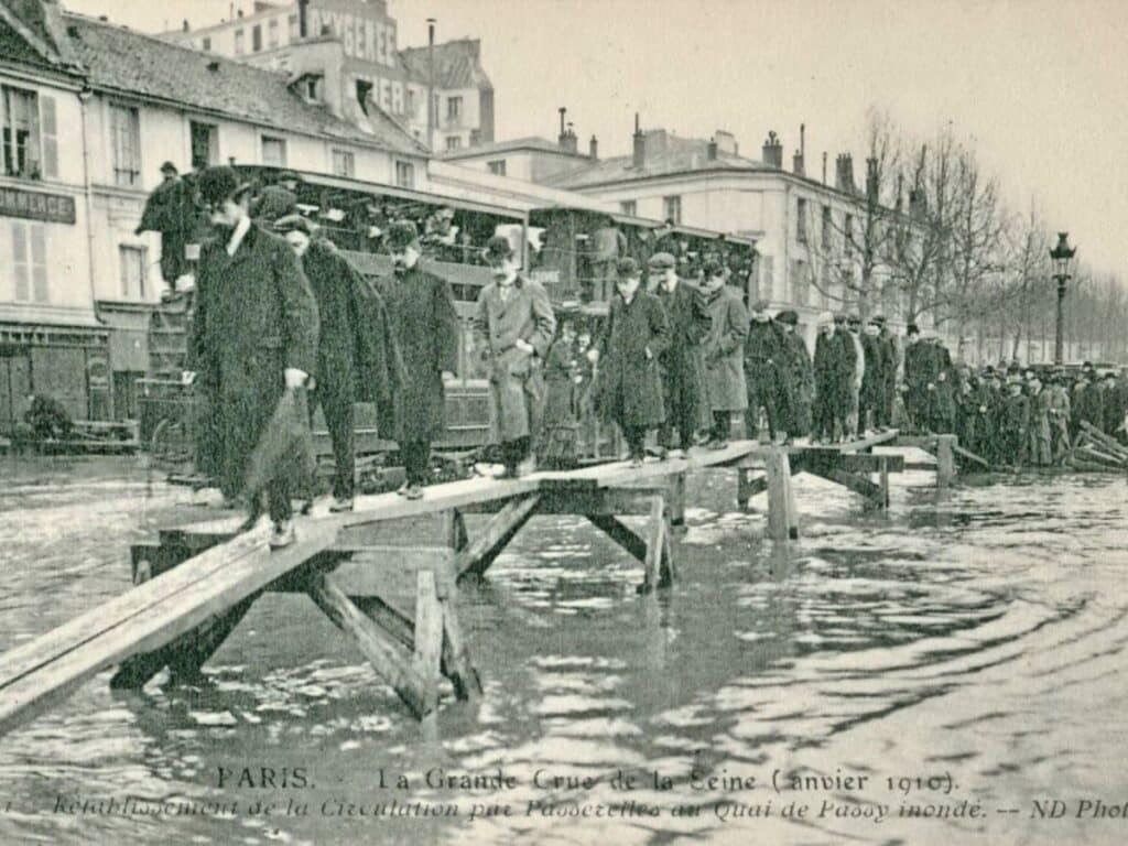 A black-and-white historical photograph showing people walking on a makeshift wooden walkway over flooded streets in Paris. The image captures the resilience of Parisians during a challenging time.