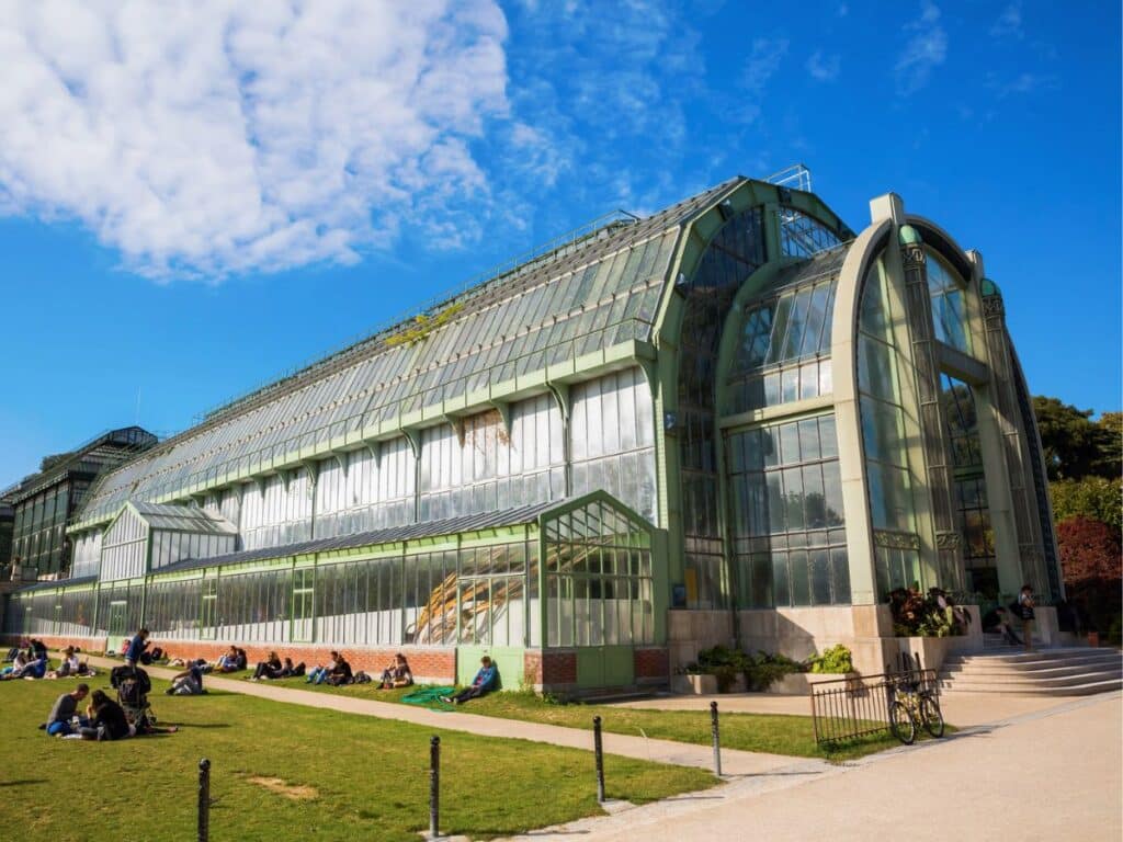 A spacious, glass-walled greenhouse in the Jardin des Plantes, surrounded by green lawns and visitors relaxing on the grass. The structure’s arched glass panels highlight its classic botanical garden design.