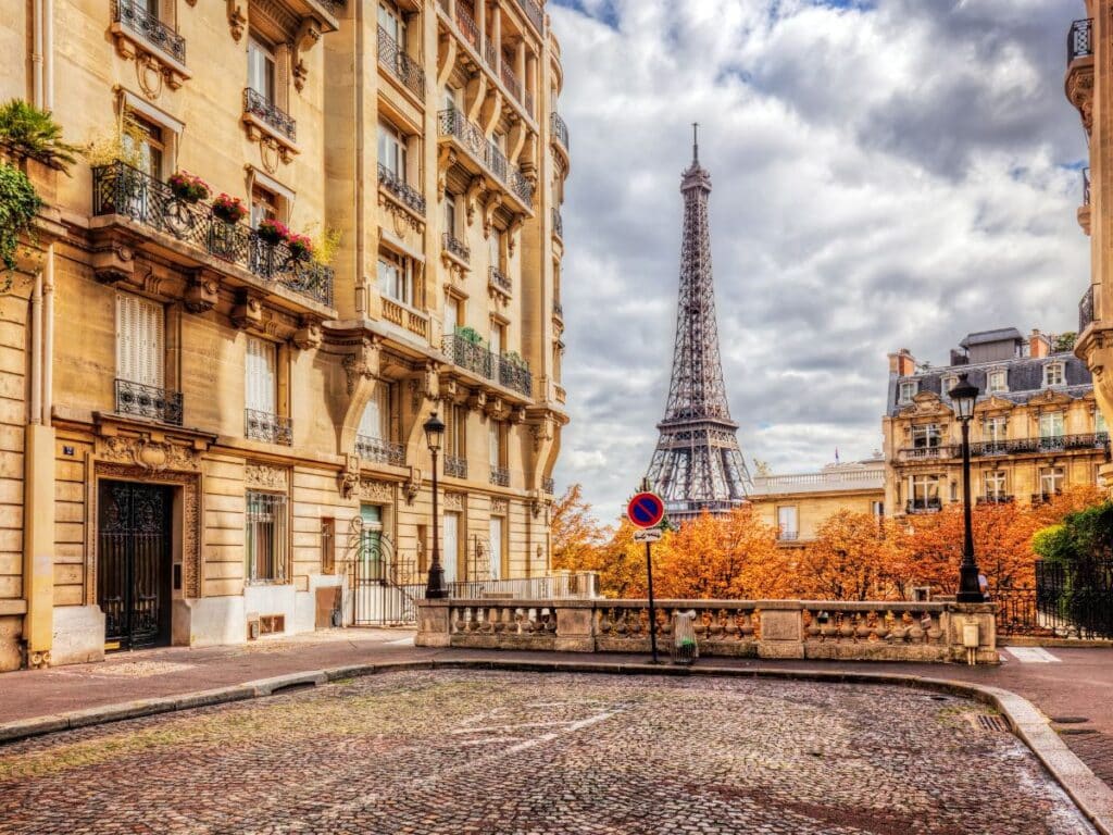 A picturesque Parisian street scene with classic French architecture and iron balconies, featuring the Eiffel Tower in the background. The autumn trees add a warm touch, with their golden leaves contrasting against the stone facades, under a partly cloudy sky.