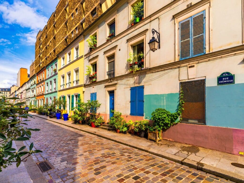 A vibrant view of Rue Crémieux in Paris, with a cobblestone street lined by pastel-colored townhouses. Each building has a unique color and is adorned with potted plants and small balconies with greenery. The street sign for Rue Crémieux is visible on the right, adding to the picturesque, charming ambiance of this beloved pedestrian-friendly street.