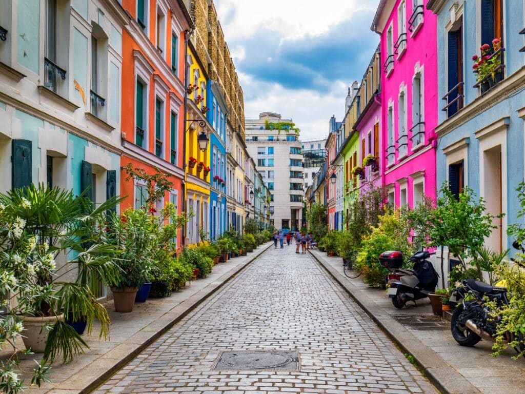 A picturesque view of Rue Crémieux with rows of colorful houses on both sides, each with unique pastel hues and decorated with potted plants. The cobblestone street adds to the quaint, village-like charm in the heart of Paris.