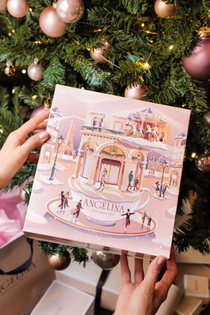 A holiday-themed Angelina gift box illustrated with Parisian landmarks and scenes, being held in front of a Christmas tree decorated with soft pink and gold ornaments.