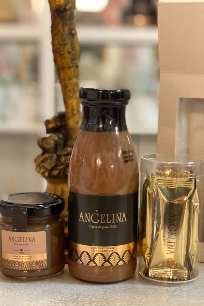 A set including a bottle of Angelina hot chocolate, a jar of chestnut cream, and a gold-wrapped Angelina chocolate, displayed on a countertop, perfect for a cozy Parisian treat.