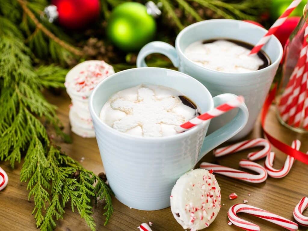 Two light blue mugs filled with hot chocolate topped with snowflake-shaped marshmallows, with candy canes and peppermint treats on the side, surrounded by festive greenery and ornaments, capturing a cozy holiday atmosphere.